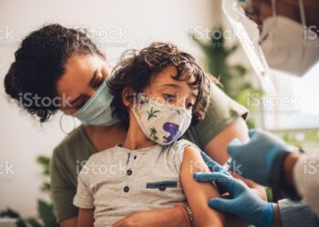 Cute boy wearing face mask taking vaccine at home. Kid with mother receiving covid vaccine from a healthcare worker at home.