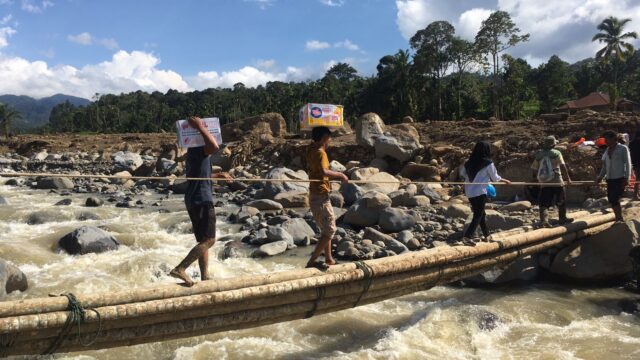 Belasan pemuda bergantian meniti batang pohon kelapa sebagai jembatan darurat yang dibentangkan di Sungai Nanggang, Jorong Subarang Aia,